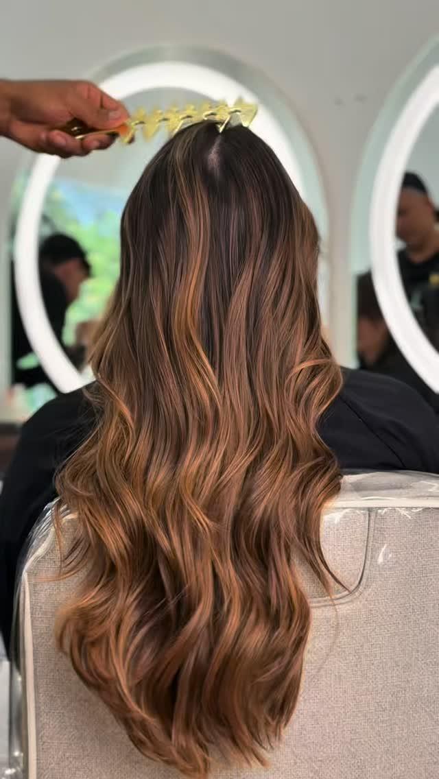 Balayage caramelo largo en ondas, de espaldas, frente a los espejos redondos del salón